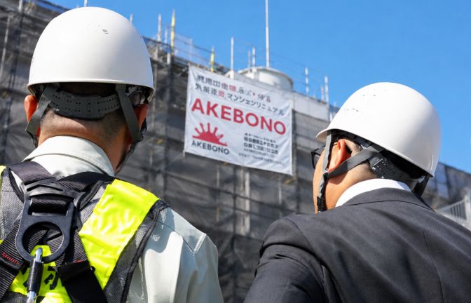 アケボノの工事看板が掛かった足場を見上げるヘルメット姿の担当者2人